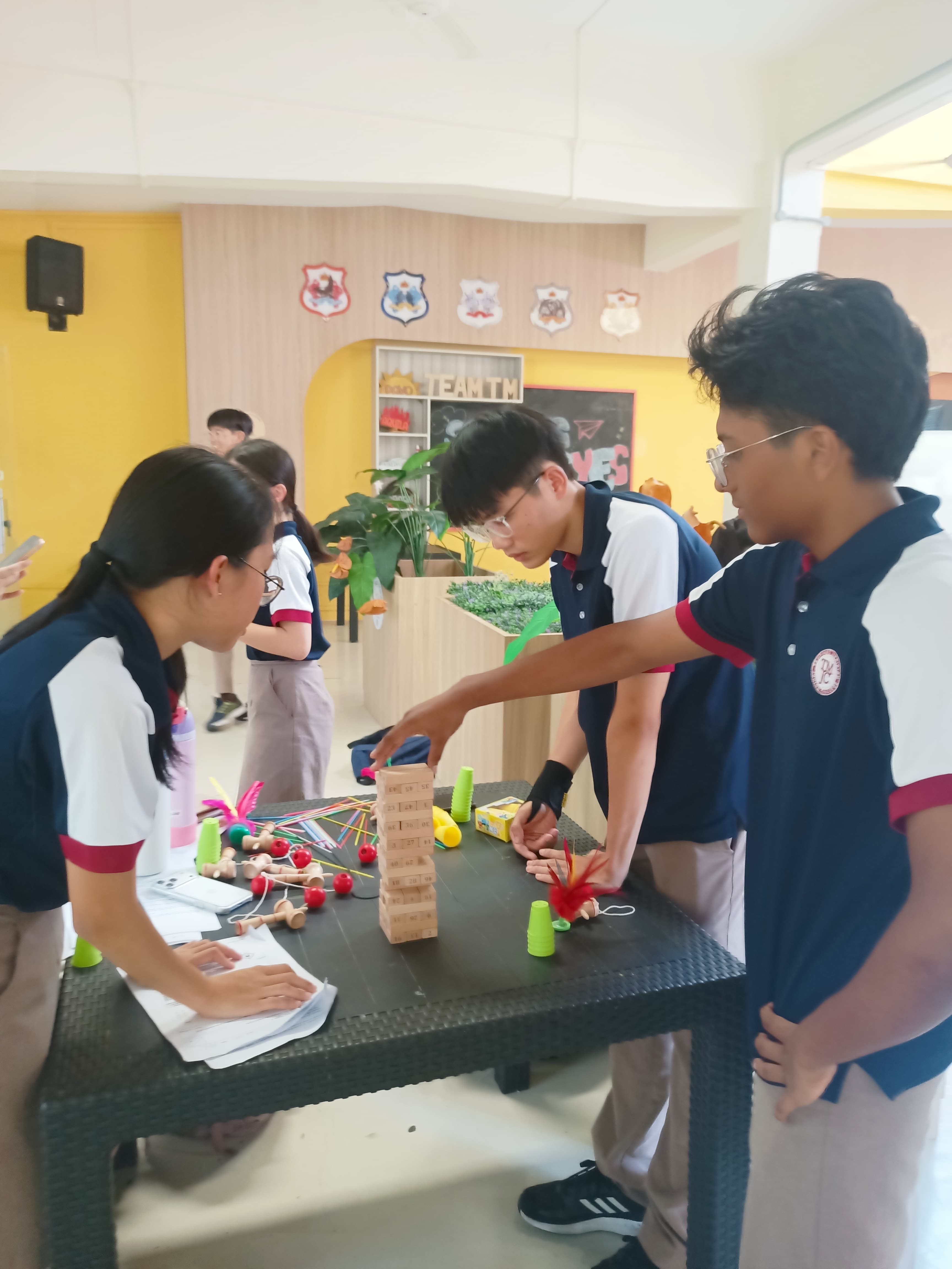 Three students in school uniforms lean over a table playing Jenga, with other games and craft materials scattered around, in a school common area.